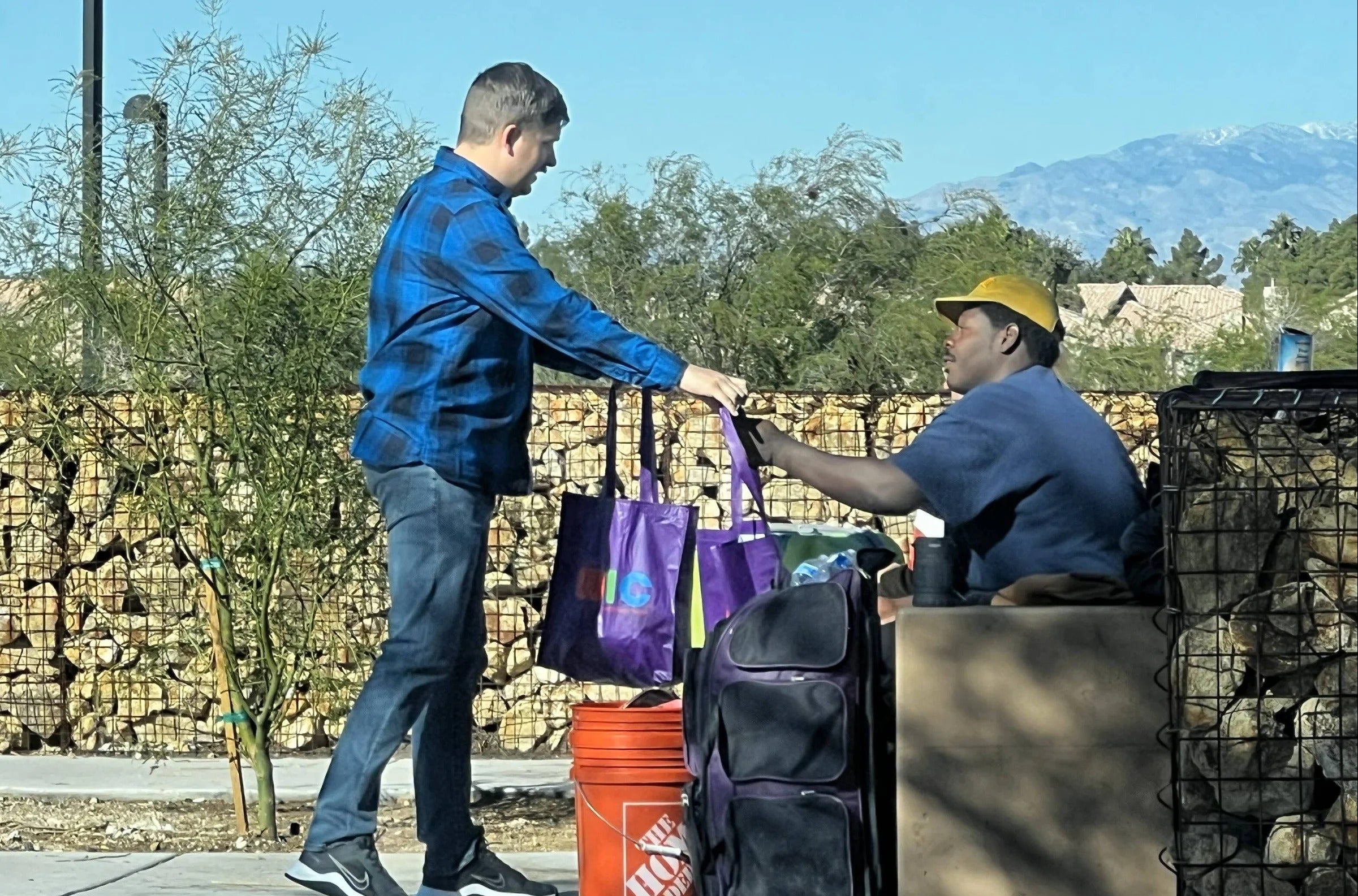 Two men interacting with bags and a bucket outdoors with mountains in the background